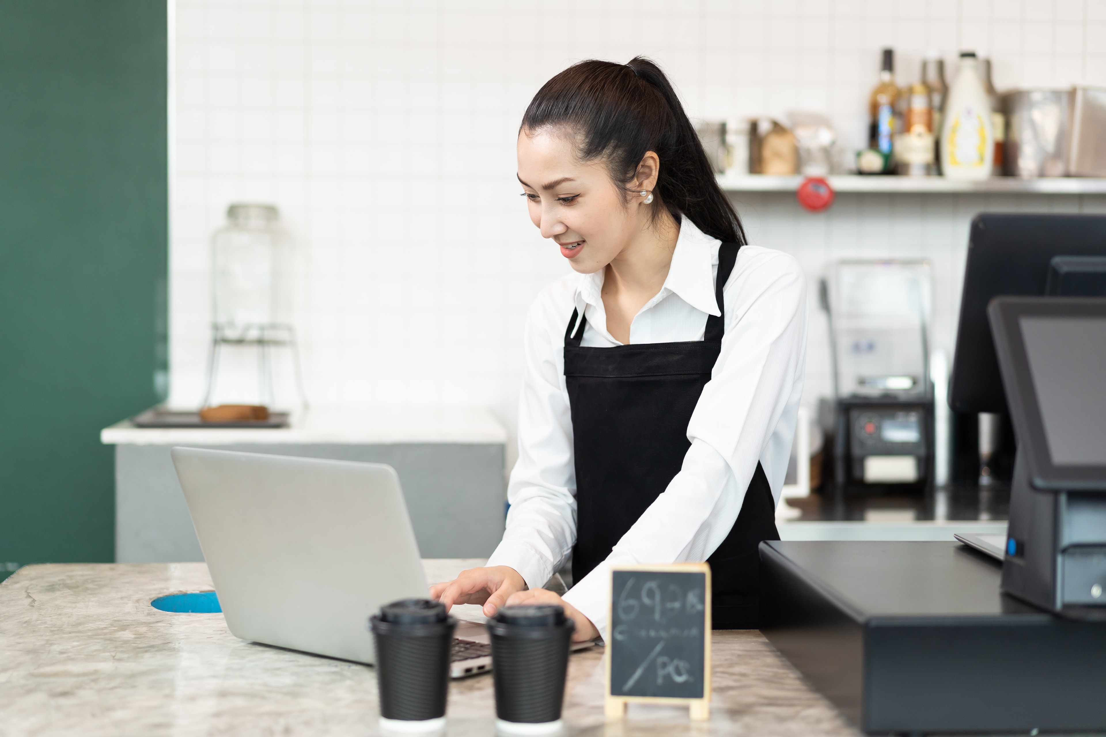 A café worker in an apron using a laptop at the counter.
