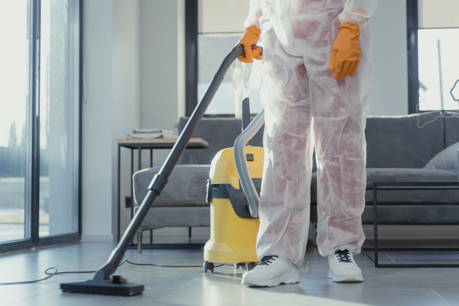 Cleaner wearing protective clothing and gloves vacuuming the floor beside a sofa.